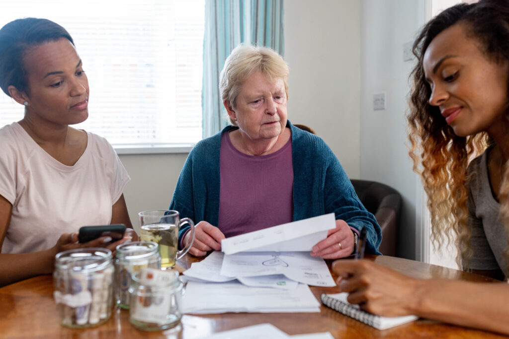 Family members go through finances with each other with uncertain expressions on their faces. The mid-female adult on the right is showing the other two something on her notepad.
